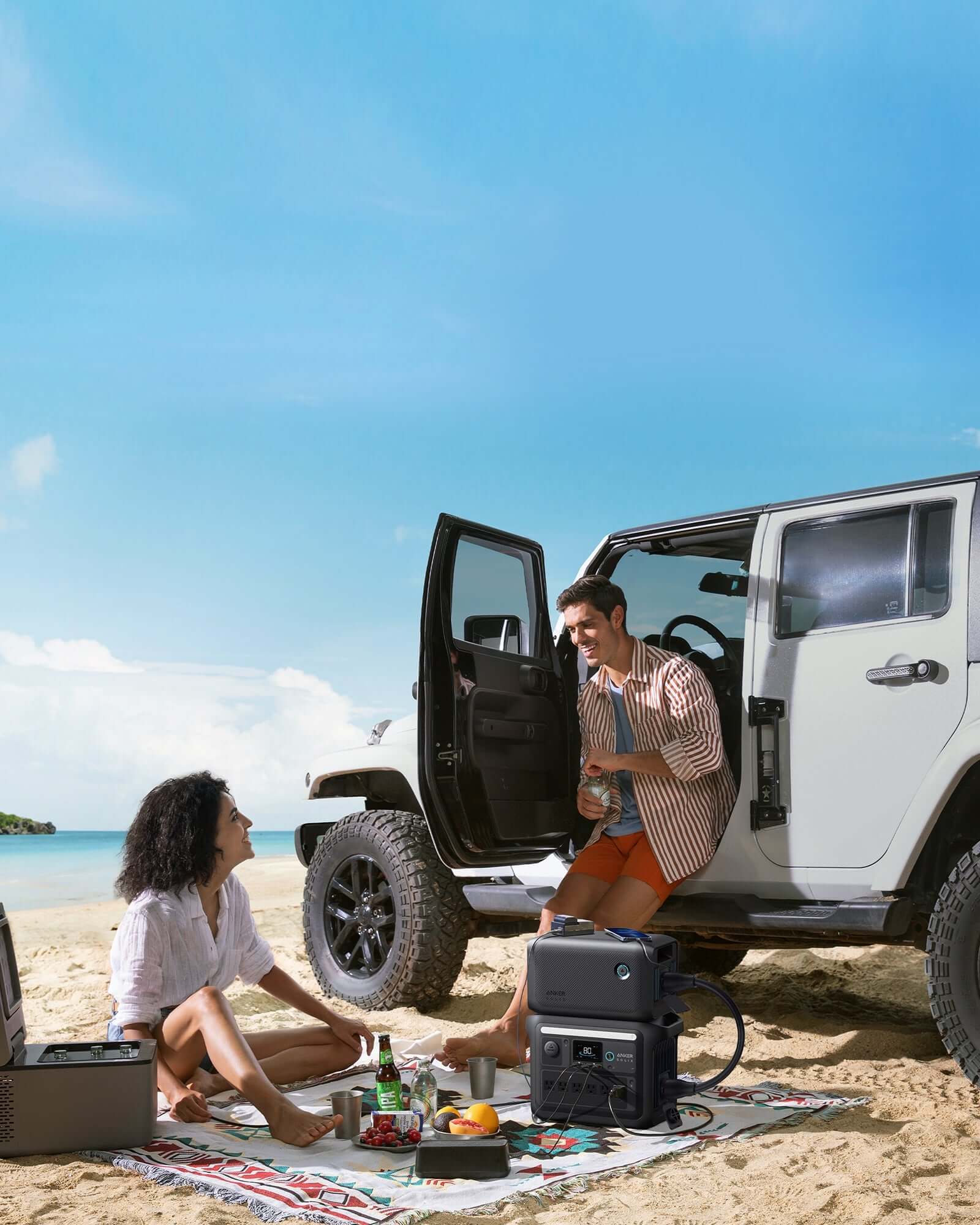 A couple enjoys a beach picnic, with the man sitting at the open door of a white SUV and the woman lounging comfortably on a blanket. Their setup is powered by an Anker SOLIX BP1000X, surrounded by snacks and drinks. The clear skies and ocean create an ideal backdrop for their seaside retreat.
