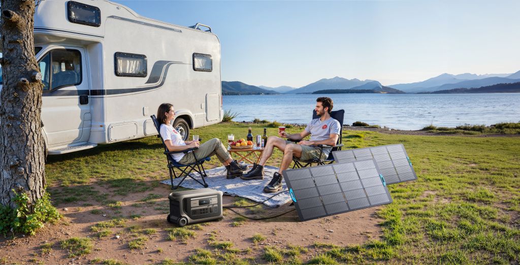 Two people relax in camping chairs by a lake near their camper van. An Anker portable solar panel and power station underscore efficiency, with food and drinks on a picnic blanket. Mountains complete the natural backdrop.