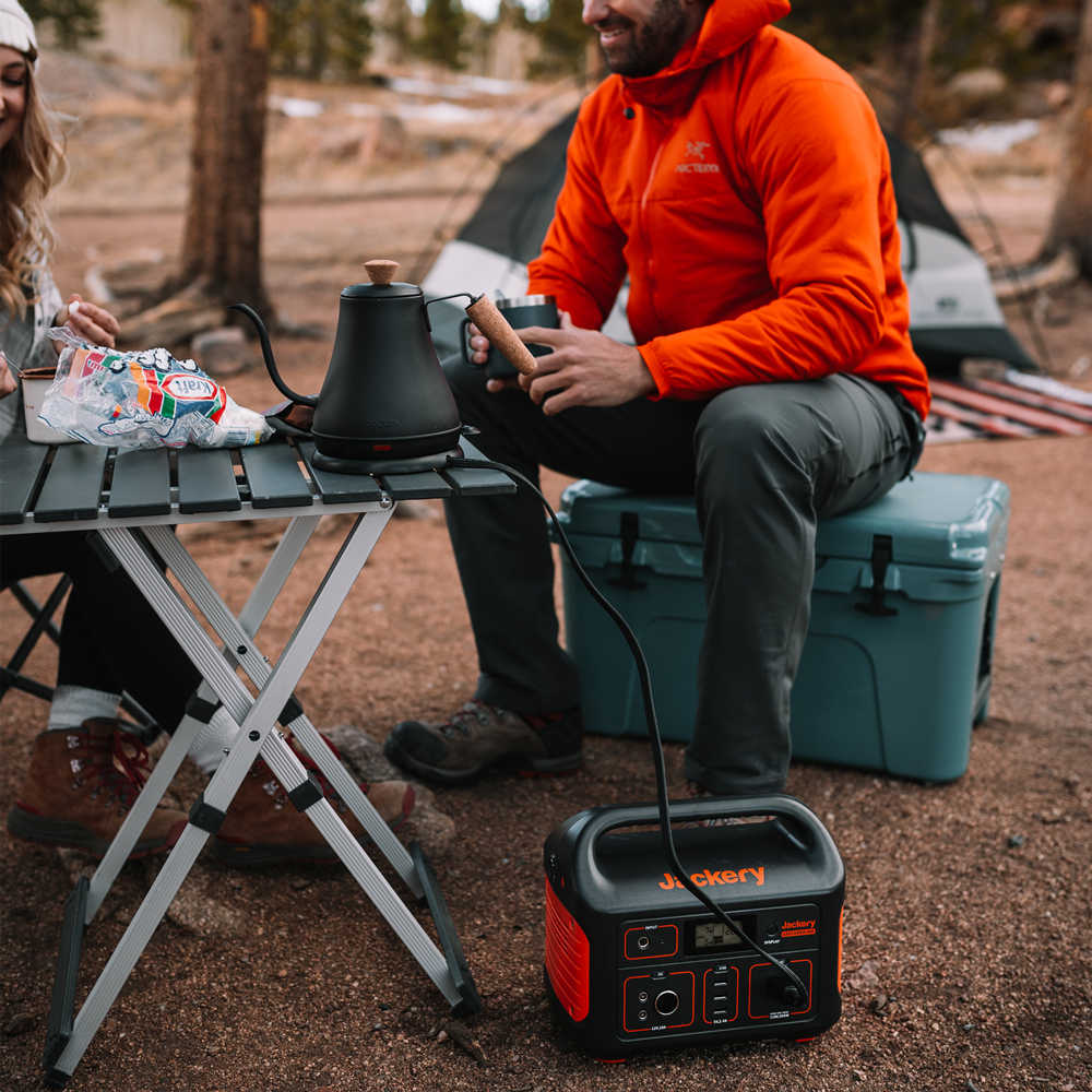 Two people relax outside near their camping tent. One sits on a Jackery portable power station with a coffee kettle, while the other has snacks at a portable table. Their reliable power station provides multiple charging options, keeping the kettle and devices powered in the great outdoors.