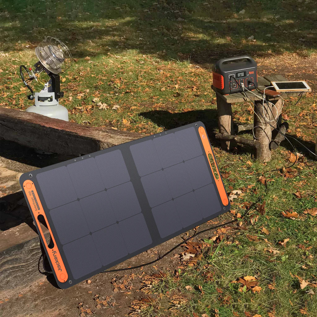 A Jackery portable solar panel is on the grass, connected to a high-capacity power station and a tablet on a wooden bench. Nearby, a small fan is hooked to a gas canister. Fallen leaves are scattered around.