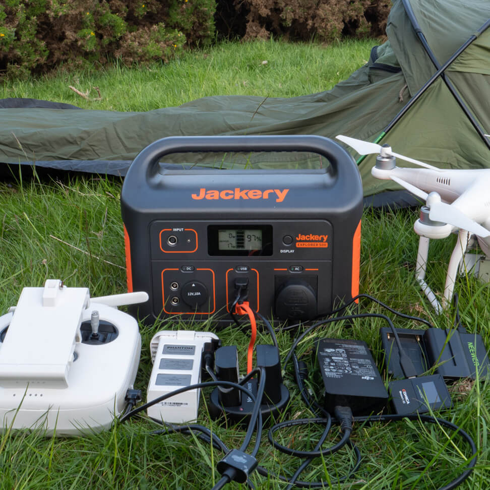 A Jackery portable power station with high battery capacity is set up on the grass next to a tent and drone, featuring various connected devices like chargers and cables, perfect for outdoor activities or camping.