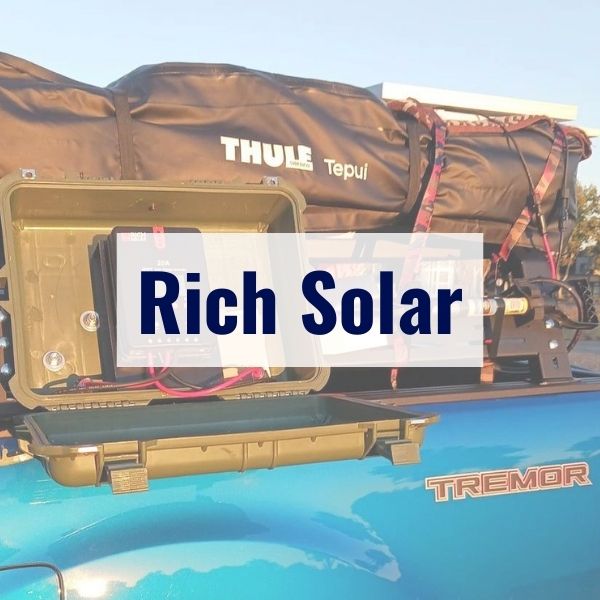 A blue truck with a bed-mounted camping setup featuring a Thule Tepui tent. The truck is equipped with a "Rich Solar" solar panel system and has a Tremor badge. The setup is designed for outdoor adventures.