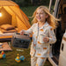 A smiling child in animal-print pajamas holds a mug and a Bluetti portable power station. A tent, van, and toys are visible outside, suggesting a camping scene. The power station’s size is labeled: 7 x 9.8 x 6.6 inches.