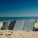 A high-efficiency Nature's Generator folding solar panel with green handles is unfolded on the sandy beach by the ocean, capturing sunlight under a clear blue sky.