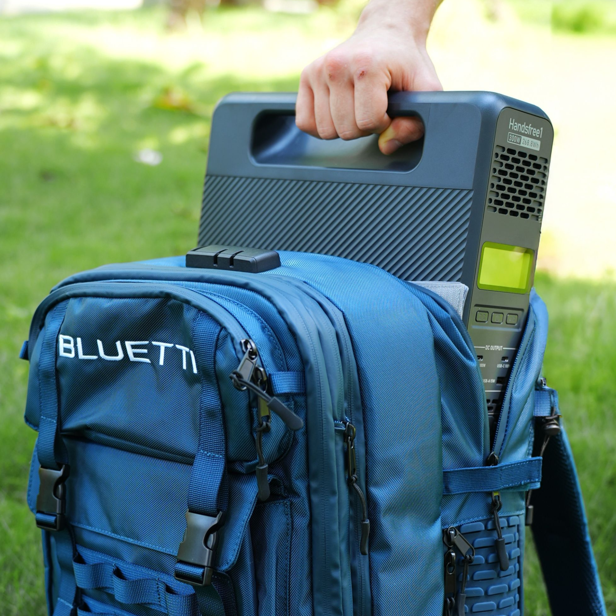 A person places a BLUETTI portable power station into a blue BLUETTI backpack on the grass outdoors, gripping the handle as the device is partially tucked inside the bag.