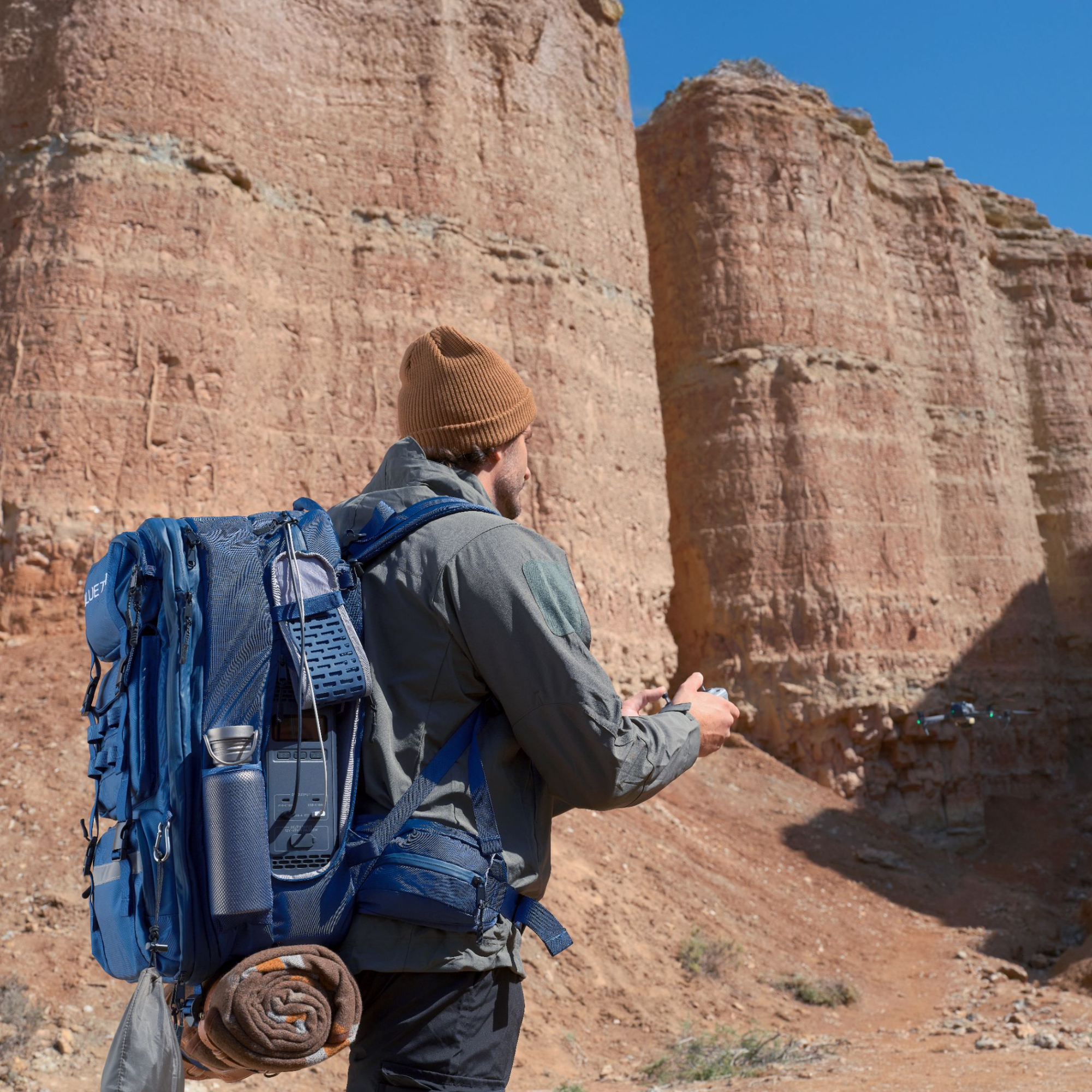 A person with a large blue Bluetti backpack and brown beanie stands facing tall, layered red rock cliffs under a clear blue sky, holding a remote controller and looking at a small drone flying nearby.