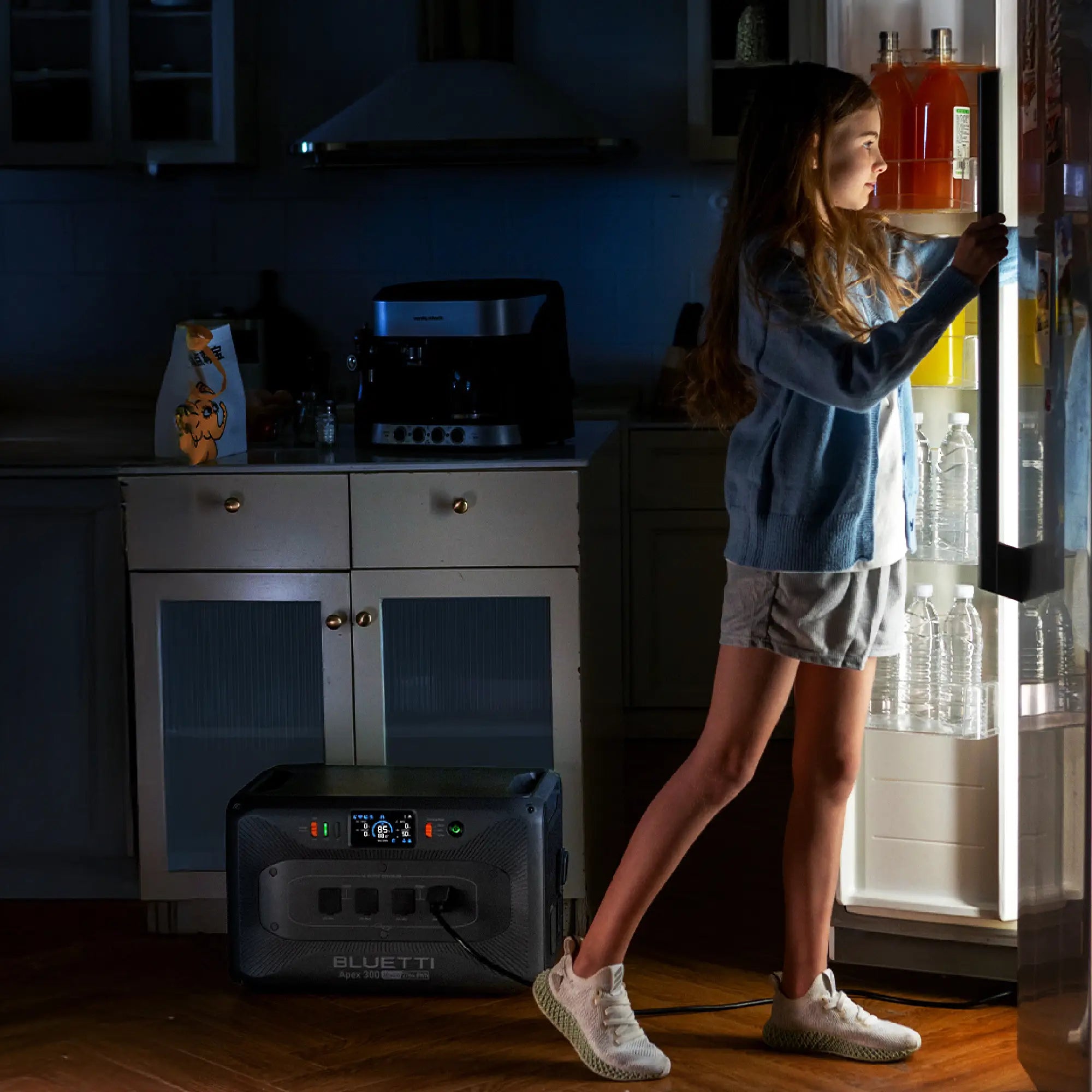 A girl in a blue sweater and shorts stands in front of an open refrigerator at night. A Bluetti portable power station is placed on the floor in the dimly lit kitchen.