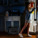 A girl in a blue sweater and shorts stands in front of an open refrigerator at night. A Bluetti portable power station is placed on the floor in the dimly lit kitchen.