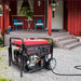 A PowerSmart 12000 watt portable generator with a red top is running on gravel in front of a red house, which has gray steps, a white door, and a patio set with two blue chairs and a round table.