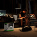 A man reads on a dark sofa in a dim room, a large dog lies nearby, and a Nature's Generator LiFePO4 portable battery for home backup sits on the floor, plugged in with its power cable.