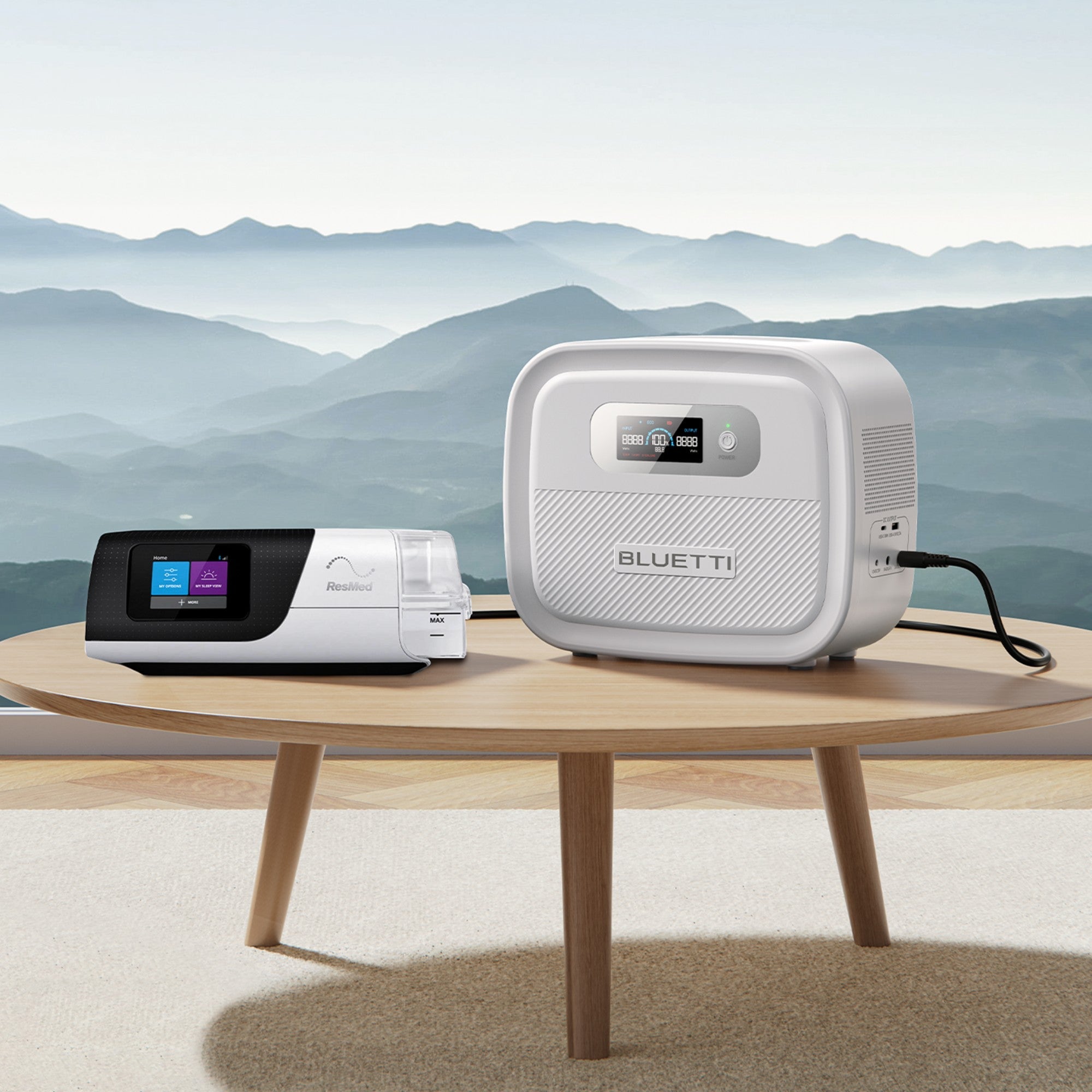 A Bluetti portable power station and a CPAP machine are placed on a round wooden table, with a scenic mountain landscape visible in the background through a large window.