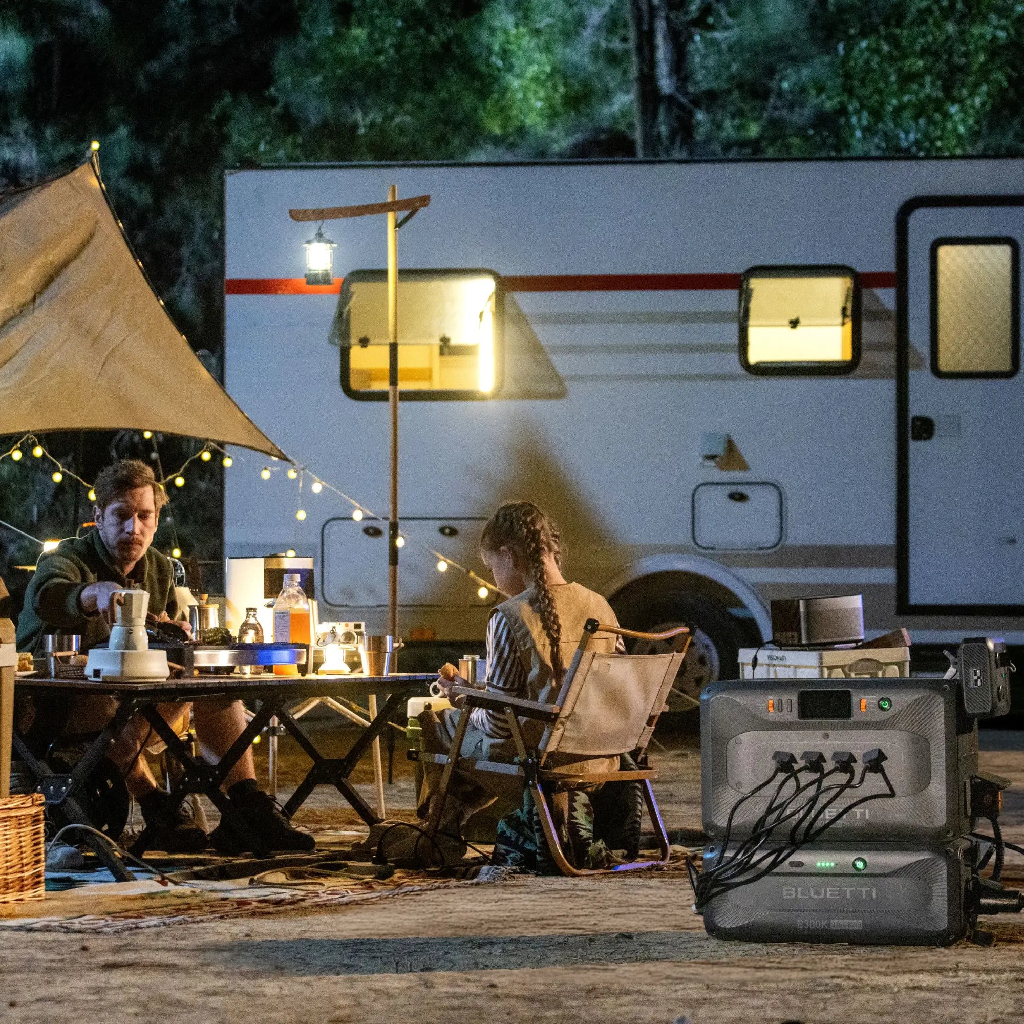 Two people sit at a table under a canopy with food and drinks, near a camper van. String lights hang overhead. In the foreground, a large Bluetti portable power station is visible. Trees are in the background.