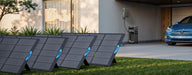 Anker solar panels are installed on a grassy lawn in front of a modern house, harnessing solar energy. A car is parked under a partially covered area beside the house, with an expandable capacity electrical unit from Anker mounted on the wall nearby.
