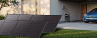 A modern home features Anker solar panels on the lawn, an expandable capacity white wall-mounted battery system, and a blue electric car in the driveway. Large windows reflect outdoor scenery.