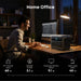 A man uses an Anker portable power station at his desk in a dimly lit room at night, powering a computer, monitor, speaker, Wi-Fi router, and desk lamp; illustrated icons and runtime details are shown.