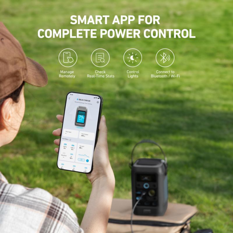 A person sits outdoors on grass, holding a smartphone with the Anker power control app open next to a portable power station. The hero section reads: manage remotely, view real-time stats, control lights, connect via Bluetooth/Wi-Fi.