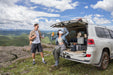 Two people with drinks stand by an SUV on rocky terrain, green hills and mountains behind them. The open trunk reveals camping and food supplies. Anker brand products are visible among the gear for outdoor convenience.