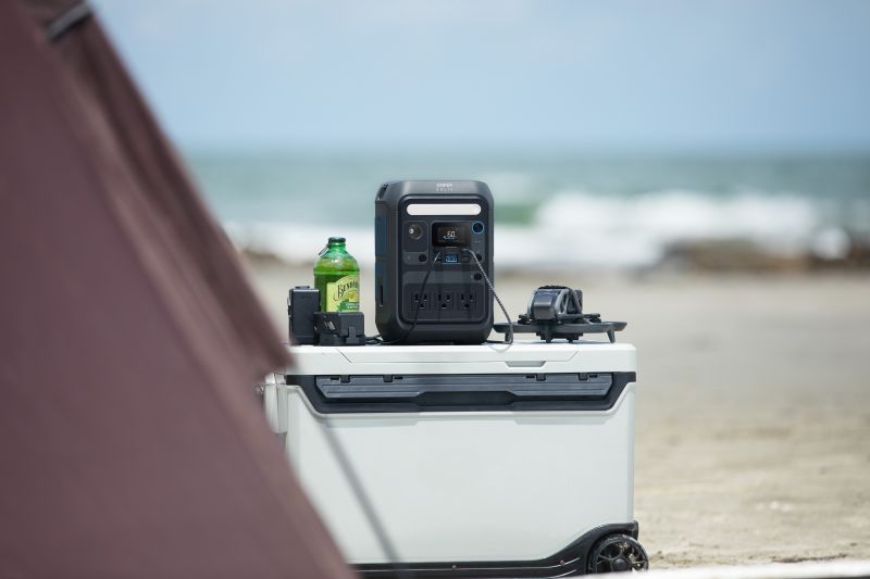 An Anker portable power station, a green bottle, and devices sit on a cooler by a tent at the beach, with the ocean and sky as a calm backdrop—an ideal outdoor setup to keep your adventures powered.