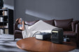 A person covered with a blanket lies on a brown sofa using a CPAP machine. In the foreground, an Anker portable power station powers the CPAP machine on a round wooden table—ideal for reliable sleep therapy anywhere.
