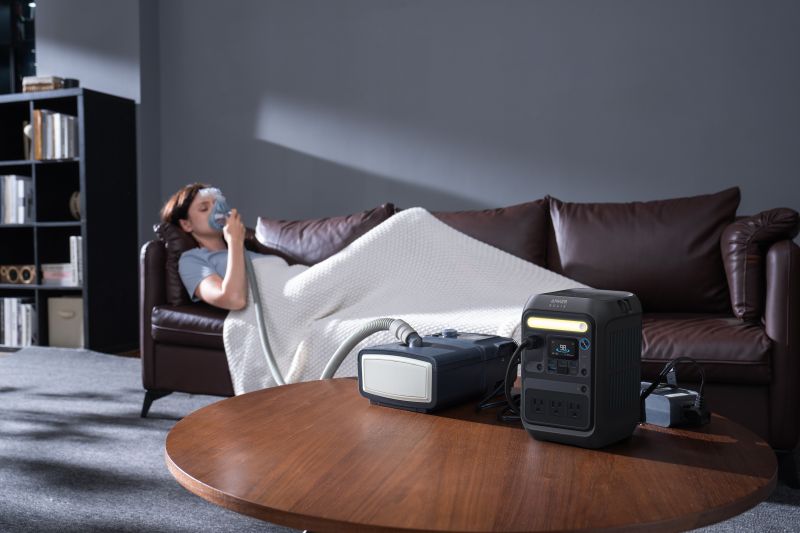 A person covered with a blanket lies on a brown sofa using a CPAP machine. In the foreground, an Anker portable power station powers the CPAP machine on a round wooden table—ideal for reliable sleep therapy anywhere.