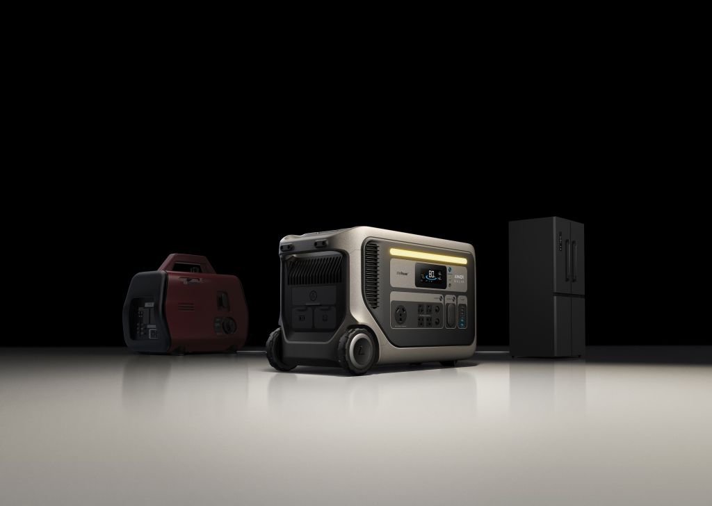 Three portable generators, including the Anker SOLIX F3000 + Expansion Battery by Anker, and a compact refrigerator are displayed on a reflective surface against a black background. The generators differ in size and style, with one centered prominently.