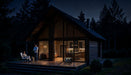 A small modern cabin with large windows glows at night, powered by an Anker solar generator. Two people are on the wooden porch—one sitting, one standing—surrounded by forest beneath a starry night sky.