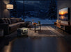 A cozy living room at night features a sofa, coffee table, large TV, and the Anker Solix F3000 solar generator on the floor. Snow falls outside sweeping window views of mountains—ideal for reliable backup power from Anker.