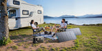 Two people relax in camping chairs by a lake near their camper van. An Anker portable solar panel and power station underscore efficiency, with food and drinks on a picnic blanket. Mountains complete the natural backdrop.