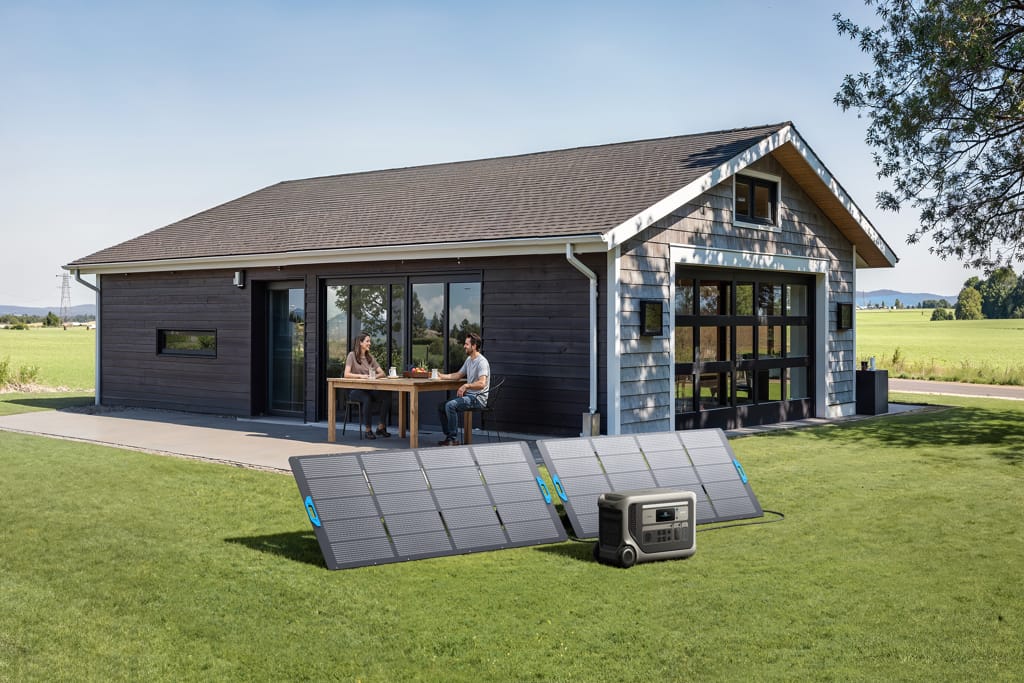 A man and woman sit at a patio table outside a modern one-story house with large windows. On the grass in front, two solar panels and an Anker Solix F3000 solar generator from Anker provide backup power.
