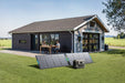 A man and woman sit at a patio table outside a modern one-story house with large windows. On the grass in front, two solar panels and an Anker Solix F3000 solar generator from Anker provide backup power.