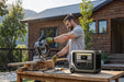 A man uses a circular saw to cut wood on a workbench outside a wooden house, with an Anker portable power station providing backup power nearby.