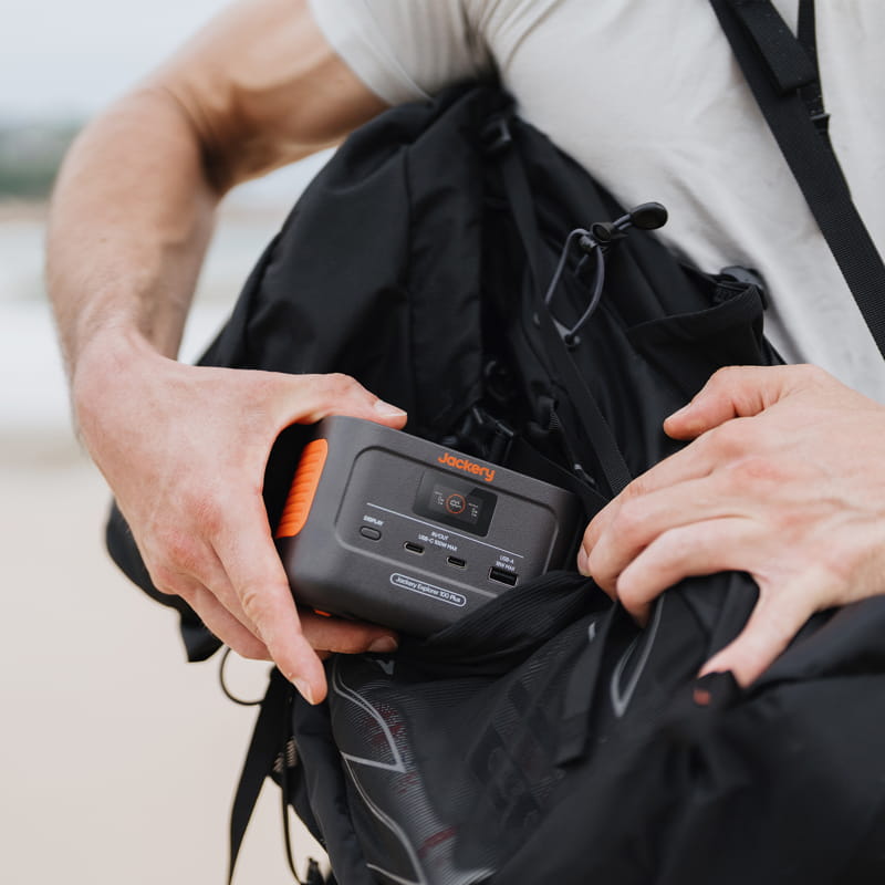 A person, wearing a white shirt with only their arms visible, places a Jackery portable power station featuring visible ports and an orange handle into a black backpack.