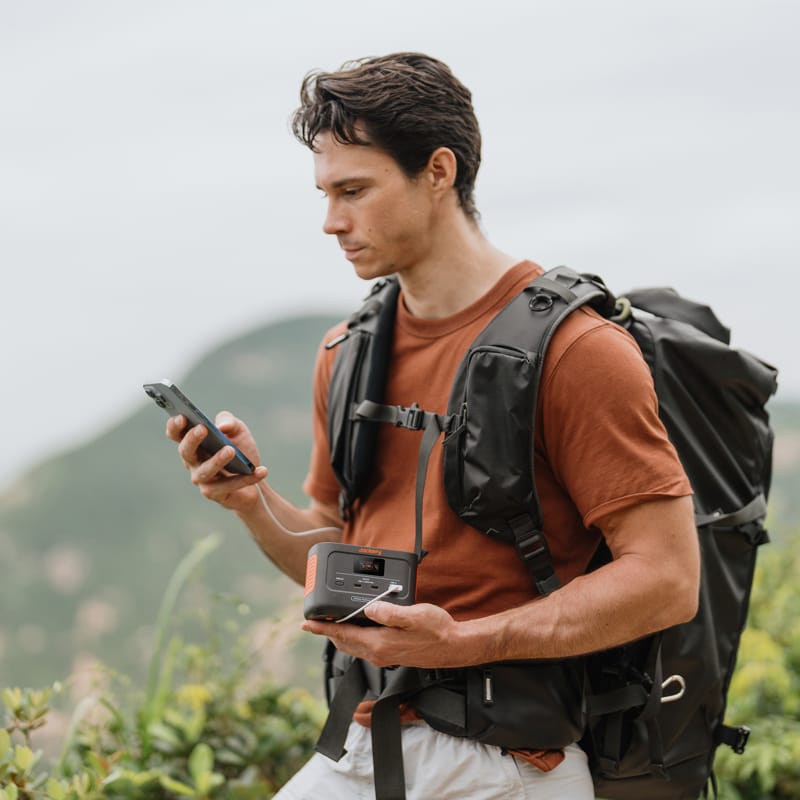 A person in an orange shirt and hiking gear stands outdoors, holding a smartphone and a Jackery portable device, with a blurred natural landscape in the background.