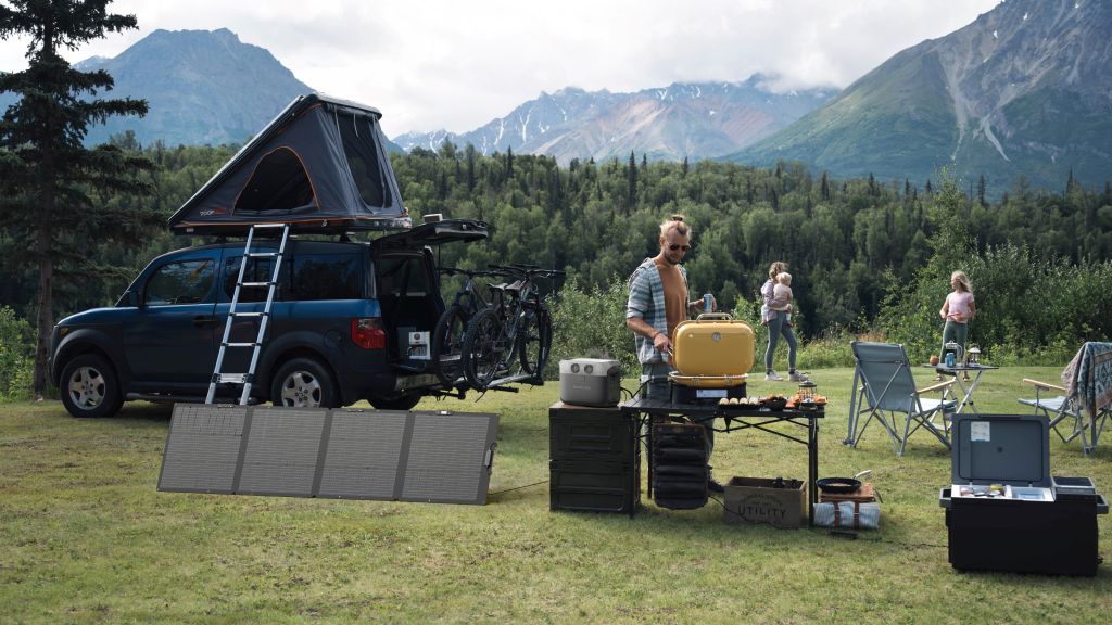A man cooks at a campsite with a yellow stove on a table, powered by the EcoFlow DELTA 3 1500 Portable Power Station. Nearby, a blue SUV with bikes and a roof tent is parked among scenic mountains and trees.