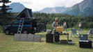 A man cooks at a campsite with a yellow stove on a table, powered by the EcoFlow DELTA 3 1500 Portable Power Station. Nearby, a blue SUV with bikes and a roof tent is parked among scenic mountains and trees.