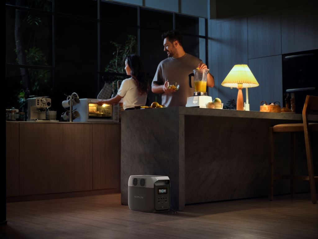 A man uses a blender at the kitchen island at night, while a woman stands by the counter. An EcoFlow portable power station with a LiFePO4 battery is on the floor near the island in their modern kitchen.