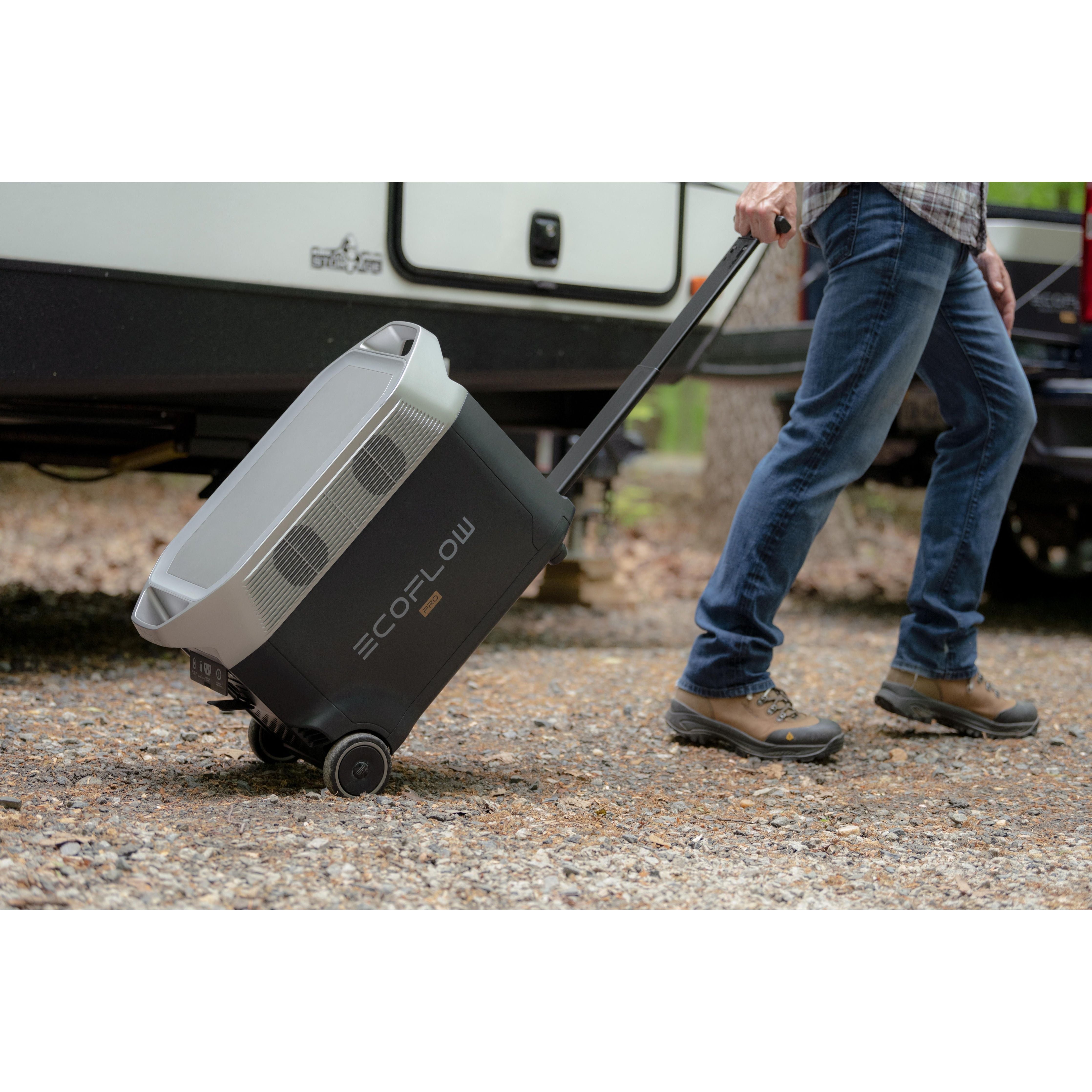 An individual in jeans and brown outdoor shoes walks across gravel, easily towing the EcoFlow DELTA Pro using its wheels and handle. This EcoFlow portable power station, equipped with X-Boost technology, is by their side next to an off-road vehicle.