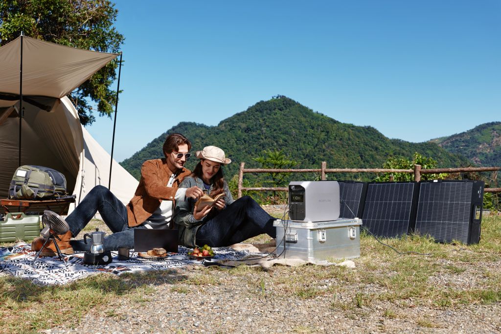 Two people enjoy a meal by their tent, with the EcoFlow DELTA 3 CLASSIC portable power station and solar panels nearby. The mountains and blue sky provide an ideal backdrop for fast outdoor charging with EcoFlow.