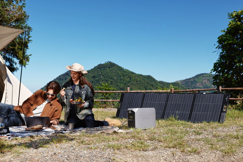 Two people relax outdoors near a tent and solar panels connected to an EcoFlow portable power station, enjoying fast charging amid green hills and a clear blue sky.