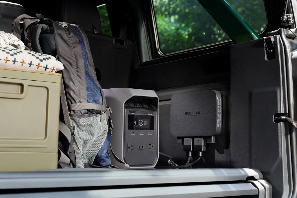 An EcoFlow portable power station and adapter sit in the back of a vehicle beside a backpack, green container, and folded blanket. Experience fast charging on the go with EcoFlow and enjoy nature views through the window.