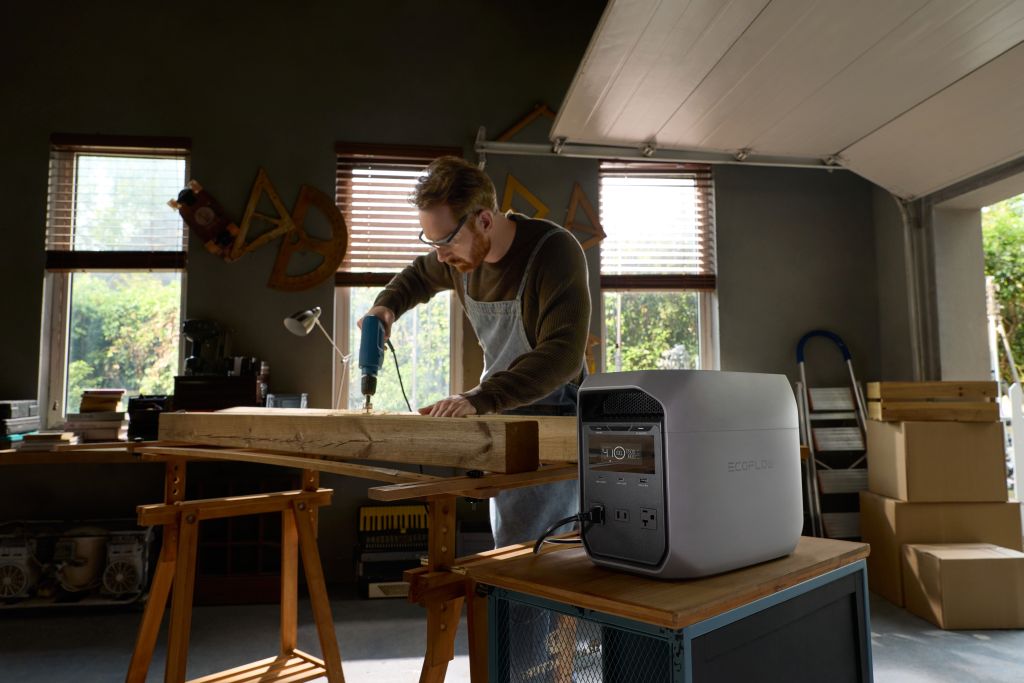 Wearing safety glasses and an apron, a person uses a power drill on wood in a workshop. On a cart nearby is an EcoFlow DELTA 3 CLASSIC portable power station with fast charging; boxes and woodworking tools appear in the background.