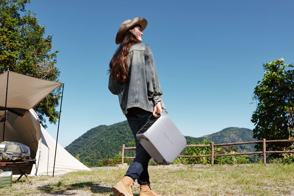 Outdoors on a sunny day, someone in a hat, denim jacket, and boots carries an EcoFlow DELTA 3 CLASSIC portable power station past camping gear and a tent, with green hills and blue sky in the background.