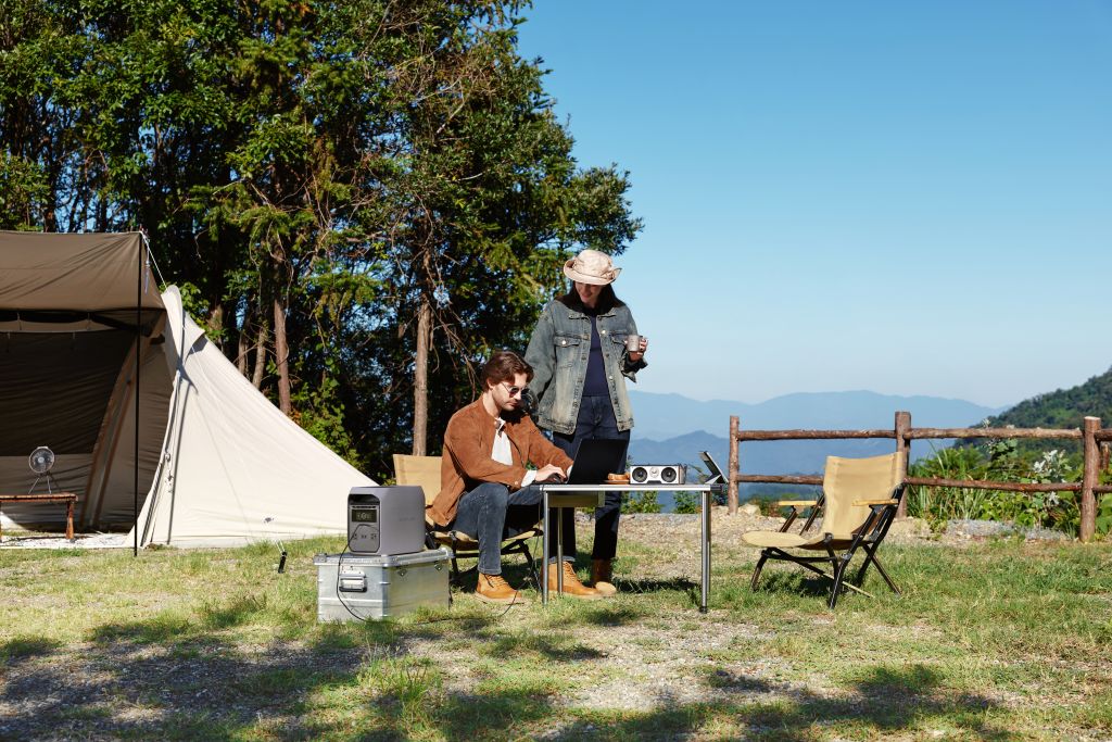At a campsite with a tent and trees, one person uses a laptop and projector powered by an EcoFlow portable power station at the table, while another stands holding a drink beneath the clear sky.