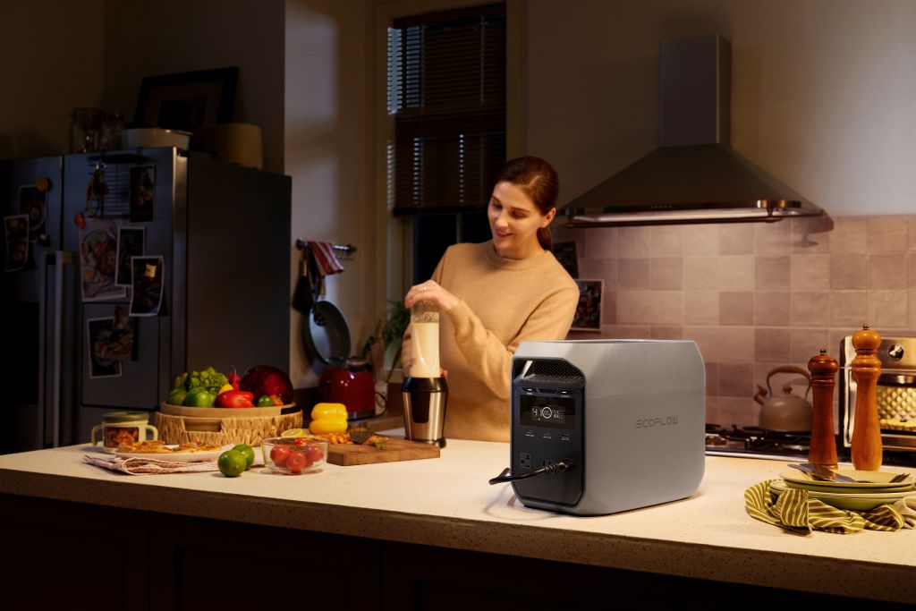 In a softly lit kitchen, a woman blends a smoothie. Fresh fruits and vegetables sit nearby, with an EcoFlow DELTA 3 CLASSIC portable power station from EcoFlow plugged into the blender for fast charging and convenience.