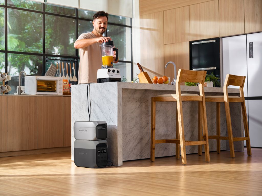A man uses a blender in a bright kitchen with wooden cabinets, plugging it into an EcoFlow backup power station on the floor during a power outage. Natural light fills the modern space as he stands at the marble island.