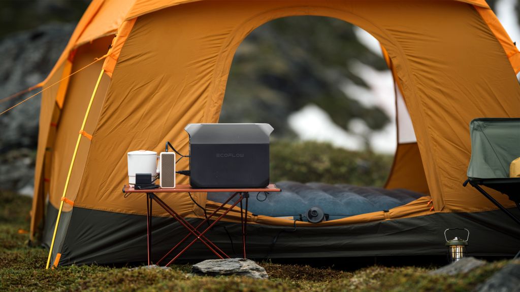 An EcoFlow portable power station, electric kettle, and smartphone sit on a foldable table outside an orange tent on grass, delivering reliable backup power for camping gear in rocky terrain—ideal for outdoor trips or power outages.