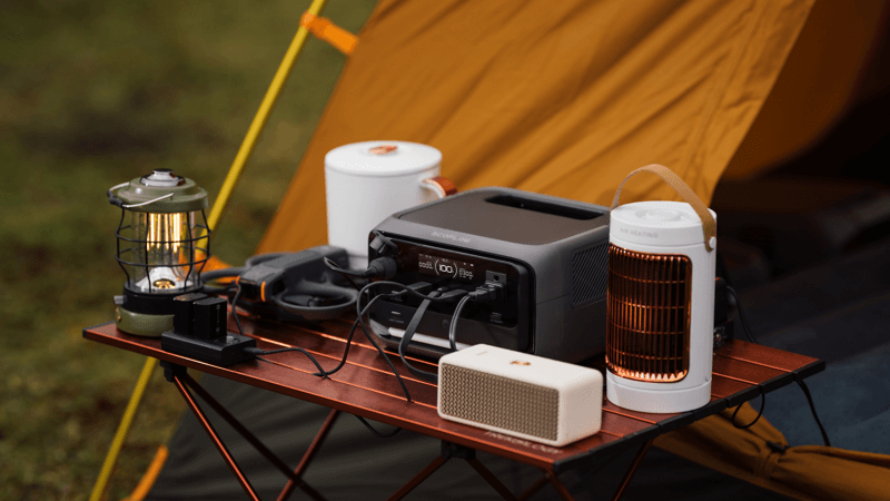 A camping setup features the EcoFlow RIVER 3 Max power station on a foldable table, amidst electronics such as a lantern, speaker, and heater; a yellow-orange tent is visible in the grassy background.