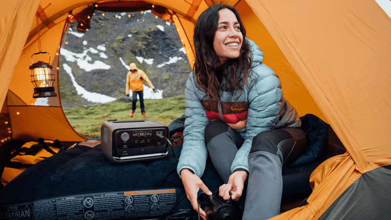 A woman in a blue jacket relaxes inside an orange tent, holding a camping lantern powered by her EcoFlow portable power station. Nearby, the unit sits beside her. In the background, someone in a yellow jacket stands on grass with snowy mountains visible.