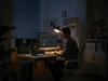 In a dimly lit room, a person uses a computer at their desk, illuminated by a lamp. Their workspace is complemented by office items and an EcoFlow portable power station from the RIVER 3 Max series, keeping them powered with its X-Boost technology for uninterrupted work.