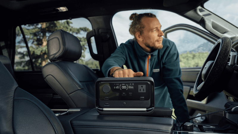 A person leans into the driver's side of a parked car, where an EcoFlow RIVER 3 Max with X-Boost technology is on the console. Its digital display shows charge levels and ports for portable power. The open car door reveals trees outside.